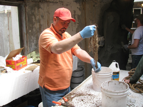 A man in an orange shirt pulls rope out from a white bucket.