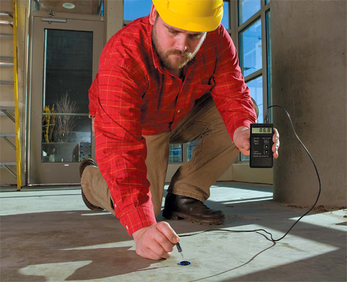 A man in a red shirt and a yellow hard hat test the concrete with a moisture meter.