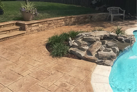 A stamped overlay surrounding an in ground pool with a faux rock feature water fall pouring into the pool.