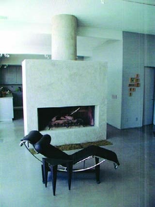 Large white modern fireplace created using a vertical concrete overlay system, stands proudly in a space between a kitchen and dining room with a black modern chrome lounge chair.