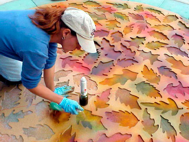 Woman spraying acrylic stain with Preval sprayer onto leaf art on a city street.