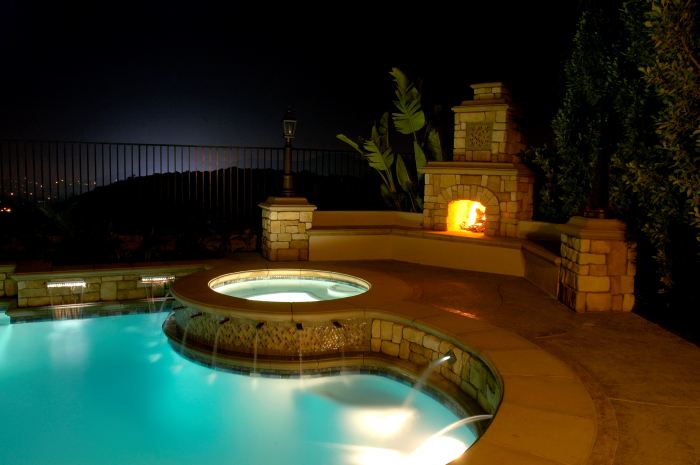 A look at a pooldeck that is light up at night with a large outdoor fireplaces in the background.