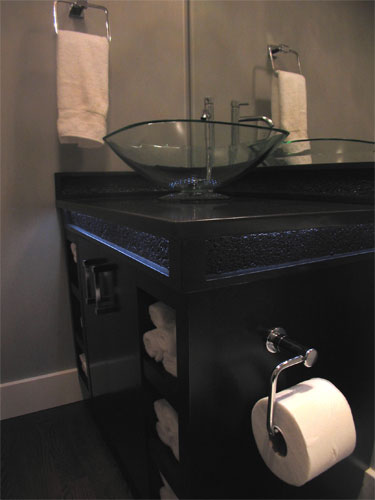 A roll of toilet paper attached to the side of this concrete vanity with a glass bowl sink.