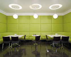 LIme green cushioned walls in a break room. Three round lights reflect circles on the ceiling above the black chairs and white tables.