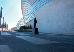 Outside a football stadium a statue stands in front of a concrete wall. The concrete patio has inset pads of flat finished concrete surrounded by a sand finish.