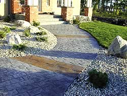 Curved stamped concrete walk way to a house. The walk way has two patterns of stamped concrete, one brick which matches the houses columns the other larger brick paver style in a lighter color.