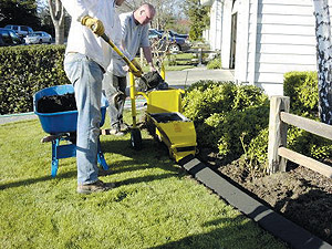 Two men using a concrete curbing machine, one is guiding the curbing machine as the other fills it with concrete.