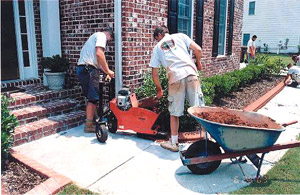 Two men and a wheel barrow filling a concrete curbing machine with integrally color concrete in a tight space.